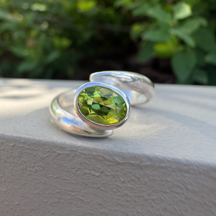 A silver ring with an oval green faceted gemstone, resting on a textured light gray surface with a blurred green garden background.