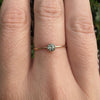 A close-up of a hand wearing a delicate gold ring with a small round green gemstone, set against a blurred green outdoor background.
