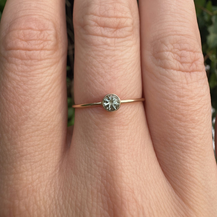 A close-up of a hand wearing a delicate gold ring with a small round green gemstone, set against a blurred green outdoor background.