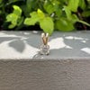 A yellow gold solitaire pendant with a clear round-cut gemstone resting on a textured gray concrete ledge with green foliage in the background.