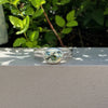 A silver ring with an oval-cut light green gemstone, resting on a textured white wall with green foliage in the background.