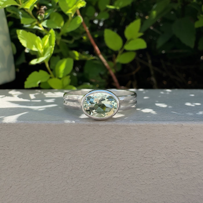 A silver ring with an oval-cut light green gemstone, resting on a textured white wall with green foliage in the background.