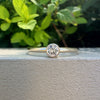 A single solitaire diamond ring in a yellow gold band, resting on a textured white surface with a blurred green garden background.
