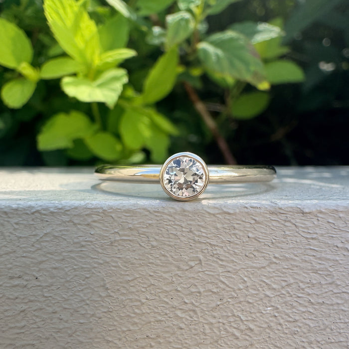 A single solitaire diamond ring in a yellow gold band, resting on a textured white surface with a blurred green garden background.
