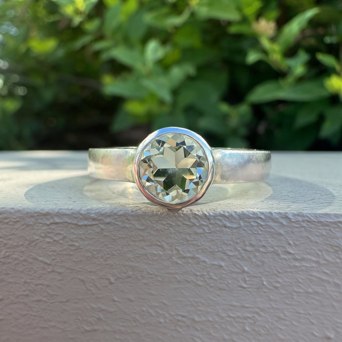 A silver ring with a large round green gemstone in a bezel setting, resting on a textured white ledge with a blurred green garden background.
