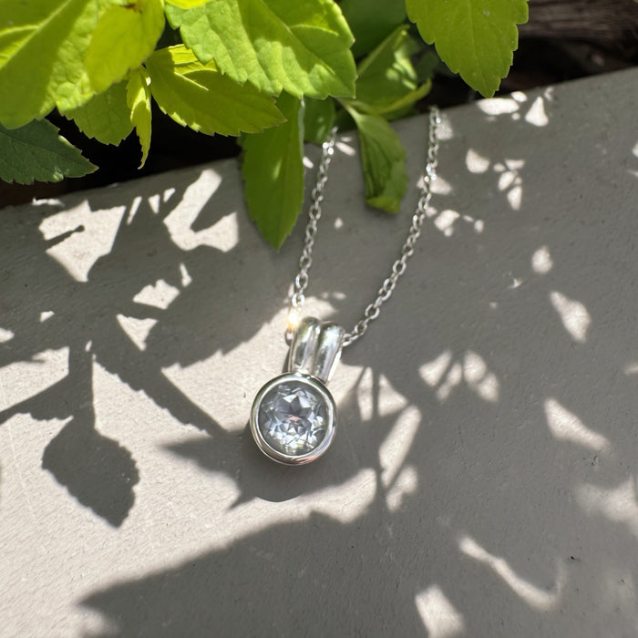 A silver pendant necklace with a clear round gemstone resting on a gray stone surface under green plant leaves and dappled sunlight.