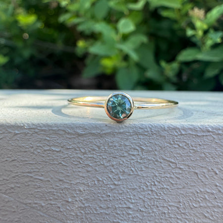 A delicate gold ring with a round bezel-set teal gemstone, resting on a textured white surface with a blurred green garden background.