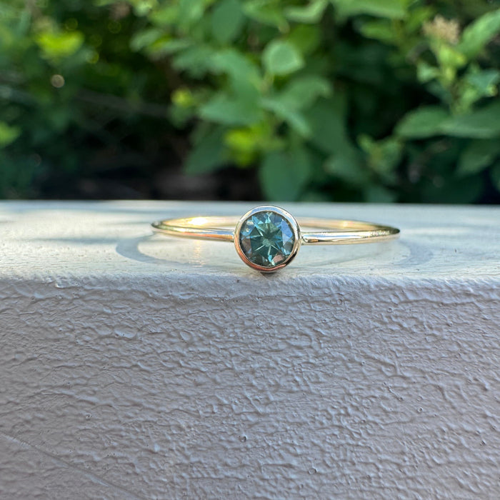 A delicate gold ring with a round bezel-set teal gemstone, resting on a textured white surface with a blurred green garden background.