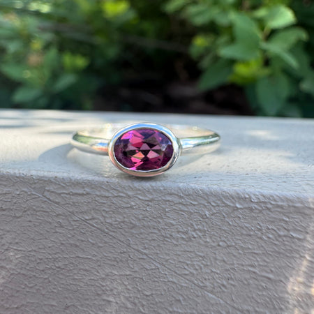 A silver ring with an oval faceted magenta-purple gemstone, resting on a textured light gray surface with a blurred green garden background.