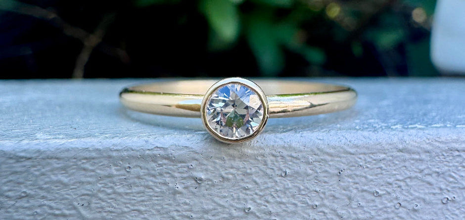 A yellow gold solitaire ring with a round-cut clear gemstone, resting on a textured white surface with a blurred green garden background.