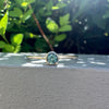 A gold ring with a round green gemstone resting on a textured gray surface, with a blurred background of green foliage and bright sunlight.