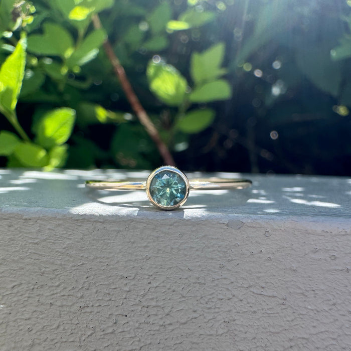 A gold ring with a round green gemstone resting on a textured gray surface, with a blurred background of green foliage and bright sunlight.
