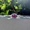 A silver ring with an oval faceted pink gemstone in a bezel setting, resting on a gray concrete ledge outdoors, with green foliage and bright sunlight in the background.