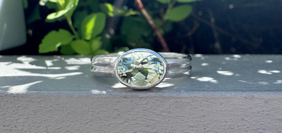 A silver ring with a oval bezel-set light blue gemstone, resting on a gray textured surface with a blurred background of green foliage and sunlight.