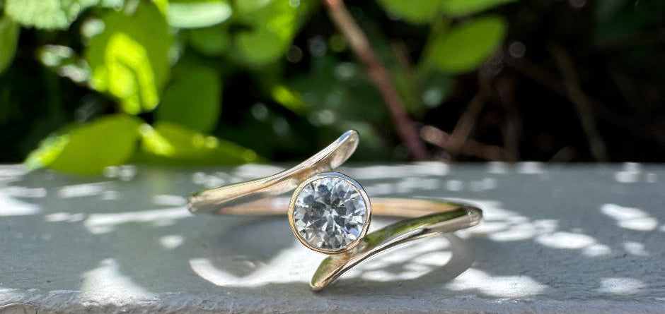 A gold ring with a round clear gemstone, resting on a textured gray surface with a blurred background of green foliage.